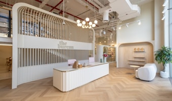 A modern reception area with a sleek, curved reception desk at the center, decorated with wicker baskets and a vase containing small decorative objects. The background features an arched structure with vertical wooden slats that house the sign 'the bureau.' To the right, a cozy seating area includes two white armchairs, a small round table, and potted plants. The ceiling is adorned with industrial-style exposed pipes and ducts, with hanging lights providing illumination. The floor is designed with a herringbone pattern.