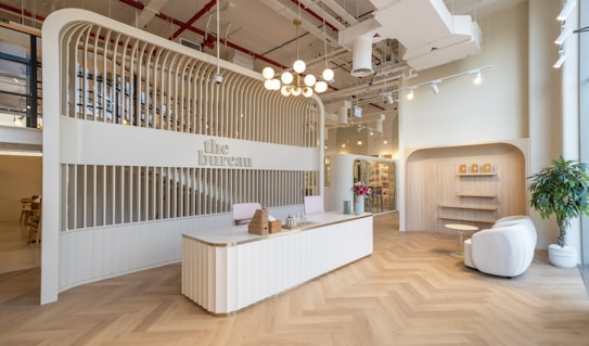 A modern reception area with a sleek, curved reception desk at the center, decorated with wicker baskets and a vase containing small decorative objects. The background features an arched structure with vertical wooden slats that house the sign 'the bureau.' To the right, a cozy seating area includes two white armchairs, a small round table, and potted plants. The ceiling is adorned with industrial-style exposed pipes and ducts, with hanging lights providing illumination. The floor is designed with a herringbone pattern.