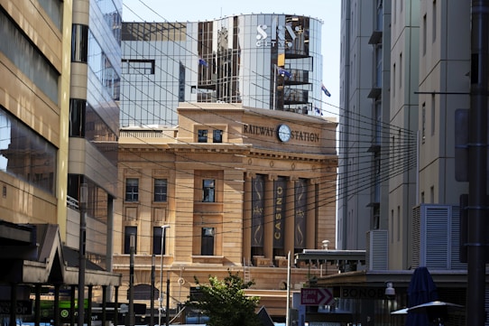 A historic building labeled 'Railway Station' is surrounded by modern high-rise architecture. The street scene includes various signs, streetlights, and overhead wires, creating a contrast between old and new architectural styles.