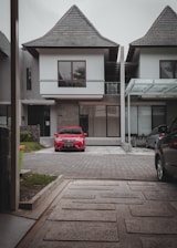 A modern two-story house featuring a steep, triangular roof and large windows stands prominently. In the driveway, a bright red car is parked, with a paved path leading from the foreground to the house. Adjacent properties can be seen, characterized by similar roof styles and contemporary design.