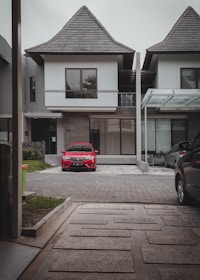 A modern two-story house featuring a steep, triangular roof and large windows stands prominently. In the driveway, a bright red car is parked, with a paved path leading from the foreground to the house. Adjacent properties can be seen, characterized by similar roof styles and contemporary design.
