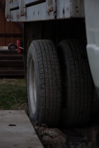 Two truck tires are positioned side by side beneath a vehicle chassis, resting on a concrete surface. The background includes a wooden structure and a red object, possibly a tool or equipment, on a platform. The setting seems to be an outdoor or garage area with patches of grass visible.