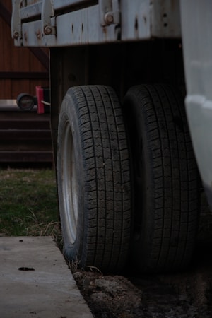 Two truck tires are positioned side by side beneath a vehicle chassis, resting on a concrete surface. The background includes a wooden structure and a red object, possibly a tool or equipment, on a platform. The setting seems to be an outdoor or garage area with patches of grass visible.