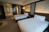 A modern hotel room with two neatly made queen-size beds featuring white sheets and pillows. The walls have an elegant design with patterned panels accompanied by soft ambient lighting. There's a subtle division between the sleeping area and a glass-enclosed shower area.