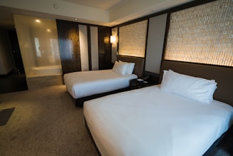 A modern hotel room with two neatly made queen-size beds featuring white sheets and pillows. The walls have an elegant design with patterned panels accompanied by soft ambient lighting. There's a subtle division between the sleeping area and a glass-enclosed shower area.