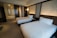 A modern hotel room with two neatly made queen-size beds featuring white sheets and pillows. The walls have an elegant design with patterned panels accompanied by soft ambient lighting. There's a subtle division between the sleeping area and a glass-enclosed shower area.