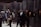 A group of people wearing headphones are engaged in viewing art or exhibits mounted on a dimly lit wall. The setting seems to be an exhibition or museum space with a dark, industrial ambiance. The viewers are standing closely to the framed artworks, which are illuminated to draw attention.