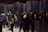 A group of people wearing headphones are engaged in viewing art or exhibits mounted on a dimly lit wall. The setting seems to be an exhibition or museum space with a dark, industrial ambiance. The viewers are standing closely to the framed artworks, which are illuminated to draw attention.