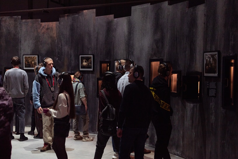 A group of people wearing headphones are engaged in viewing art or exhibits mounted on a dimly lit wall. The setting seems to be an exhibition or museum space with a dark, industrial ambiance. The viewers are standing closely to the framed artworks, which are illuminated to draw attention.