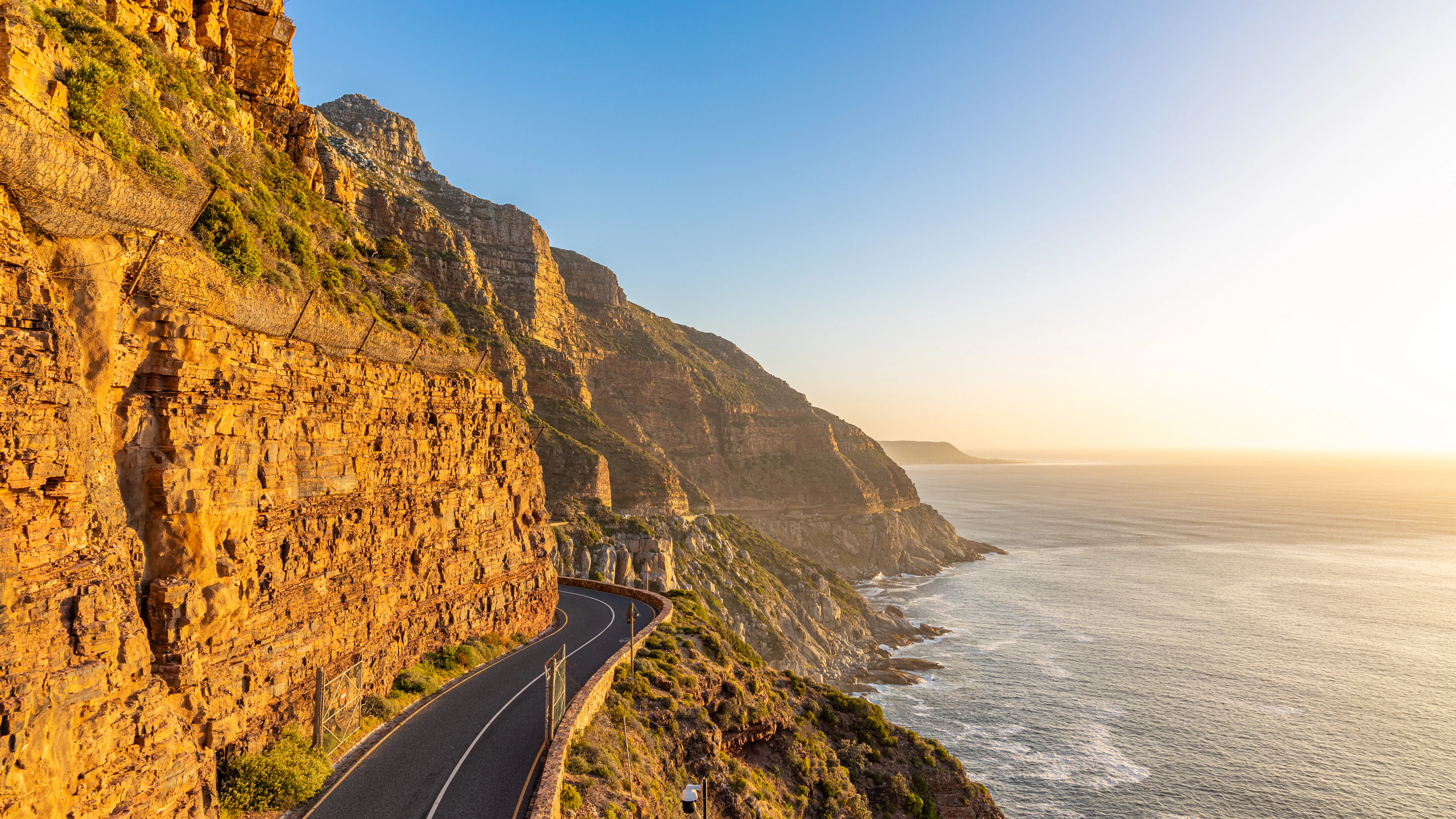 Chapman's Peak Drive curving along the golden sandstone cliff face above the Atlantic on the Cape Peninsula, the road empty in low evening sunlight and the sea glowing to the horizon