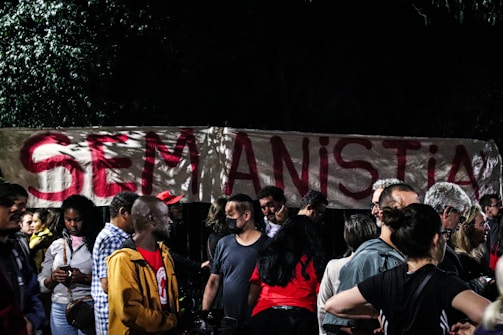 A group of people gathered in front of a large banner with the phrase 'SEM ANISTIA' written in bold red letters. The setting appears to be nighttime, with trees partially visible in the background.