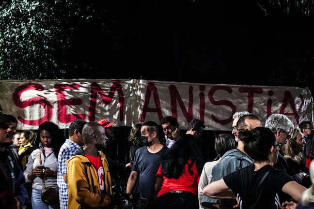 A group of people gathered in front of a large banner with the phrase 'SEM ANISTIA' written in bold red letters. The setting appears to be nighttime, with trees partially visible in the background.