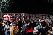 A group of people gathered in front of a large banner with the phrase 'SEM ANISTIA' written in bold red letters. The setting appears to be nighttime, with trees partially visible in the background.