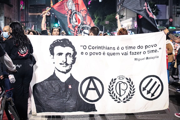 A group of people is holding a large banner featuring a black-and-white portrait of a man and several logos or symbols. The banner also displays a quote in Portuguese attributed to Miguel Bataglia. The scene takes place in a bustling urban environment with additional banners and flags being held aloft by the crowd. Some individuals are wearing masks while others are in casual attire.