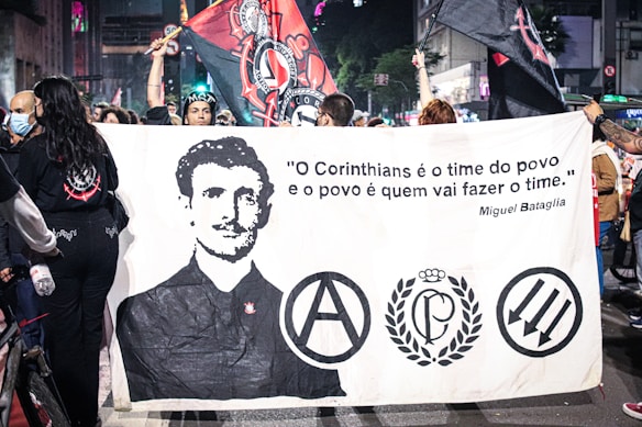 A group of people is holding a large banner featuring a black-and-white portrait of a man and several logos or symbols. The banner also displays a quote in Portuguese attributed to Miguel Bataglia. The scene takes place in a bustling urban environment with additional banners and flags being held aloft by the crowd. Some individuals are wearing masks while others are in casual attire.