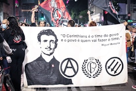 A group of people is holding a large banner featuring a black-and-white portrait of a man and several logos or symbols. The banner also displays a quote in Portuguese attributed to Miguel Bataglia. The scene takes place in a bustling urban environment with additional banners and flags being held aloft by the crowd. Some individuals are wearing masks while others are in casual attire.