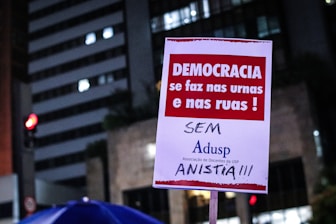 A poster is being held up at a protest. It contains Portuguese text advocating for democracy and rejecting amnesty. The background shows a cityscape with lit buildings and a traffic light.