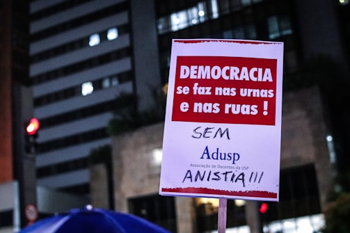 A poster is being held up at a protest. It contains Portuguese text advocating for democracy and rejecting amnesty. The background shows a cityscape with lit buildings and a traffic light.