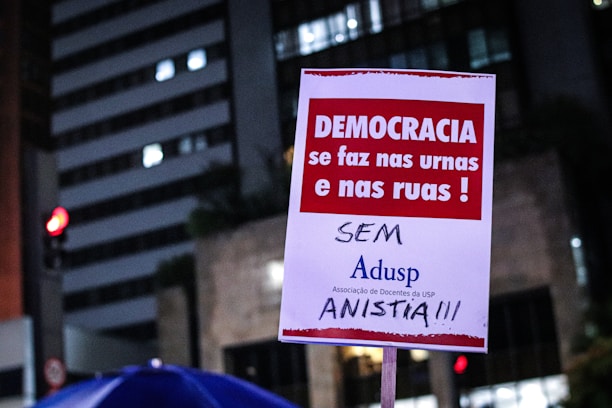 A poster is being held up at a protest. It contains Portuguese text advocating for democracy and rejecting amnesty. The background shows a cityscape with lit buildings and a traffic light.