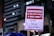 A poster is being held up at a protest. It contains Portuguese text advocating for democracy and rejecting amnesty. The background shows a cityscape with lit buildings and a traffic light.