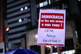 A poster is being held up at a protest. It contains Portuguese text advocating for democracy and rejecting amnesty. The background shows a cityscape with lit buildings and a traffic light.