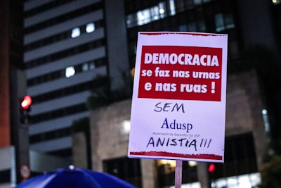 A poster is being held up at a protest. It contains Portuguese text advocating for democracy and rejecting amnesty. The background shows a cityscape with lit buildings and a traffic light.
