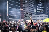 A large crowd gathers on a city street during nighttime, surrounded by tall buildings. Some people hold signs, including one with the words 'SEM ANISTIA'. There is a smoky atmosphere, and the crowd appears engaged and possibly protesting.