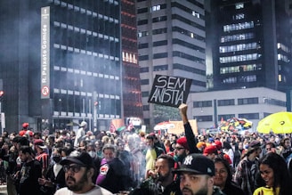A large crowd gathers on a city street during nighttime, surrounded by tall buildings. Some people hold signs, including one with the words 'SEM ANISTIA'. There is a smoky atmosphere, and the crowd appears engaged and possibly protesting.