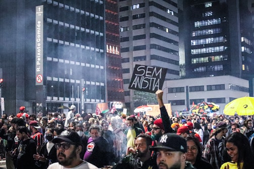 A large crowd gathers on a city street during nighttime, surrounded by tall buildings. Some people hold signs, including one with the words 'SEM ANISTIA'. There is a smoky atmosphere, and the crowd appears engaged and possibly protesting.