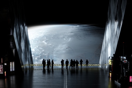 A group of people stands inside a futuristic building, looking out through large windows at a vast view of a planet. The atmosphere is mysterious, with low lighting inside emphasizing the glowing, curved surface of the planet outside. The interior has a sleek, metallic design with some illuminated panels and vending machines along the walls.