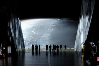 A group of people stands inside a futuristic building, looking out through large windows at a vast view of a planet. The atmosphere is mysterious, with low lighting inside emphasizing the glowing, curved surface of the planet outside. The interior has a sleek, metallic design with some illuminated panels and vending machines along the walls.