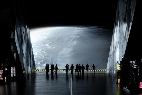 A group of people stands inside a futuristic building, looking out through large windows at a vast view of a planet. The atmosphere is mysterious, with low lighting inside emphasizing the glowing, curved surface of the planet outside. The interior has a sleek, metallic design with some illuminated panels and vending machines along the walls.