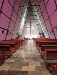 A triangular-shaped modern church interior with wooden pews arranged in rows on either side of a central aisle. At the end of the aisle, a large crucifix is mounted on the back wall above the altar. The walls are composed of large, geometric panels, and stained glass windows flank the sides near the altar.