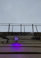A structure with metal bars extending upward toward an overcast sky. Below, a circular LED light fixture emits bright blue and red colors, casting a purple glow on the wall. A security camera is also visible, pointing outward.