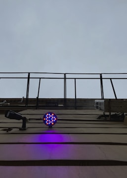 A structure with metal bars extending upward toward an overcast sky. Below, a circular LED light fixture emits bright blue and red colors, casting a purple glow on the wall. A security camera is also visible, pointing outward.