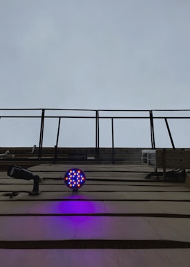 A structure with metal bars extending upward toward an overcast sky. Below, a circular LED light fixture emits bright blue and red colors, casting a purple glow on the wall. A security camera is also visible, pointing outward.