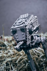 A detailed LEGO model of a futuristic machine with mechanical legs and a robust body. The model is primarily grey and appears to be a robotic walker. It is placed in an outdoor setting on a bed of dried grass or plants, with a blurred background.