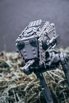 A detailed LEGO model of a futuristic machine with mechanical legs and a robust body. The model is primarily grey and appears to be a robotic walker. It is placed in an outdoor setting on a bed of dried grass or plants, with a blurred background.
