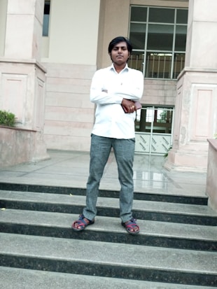 A man stands confidently on the steps in front of a building. He is wearing a white shirt, grey pants, and sandals. His arms are crossed as he looks towards the camera. The background features architectural elements, including large pillars and a glass door.