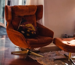 An orange-brown armchair with a matching footrest on a wooden floor. The chair features a patterned cushion with geometric designs in yellow and dark red. A light-colored rug is partially visible underneath, and sheer white curtains are drawn aside a sliding glass door, allowing daylight to filter in.