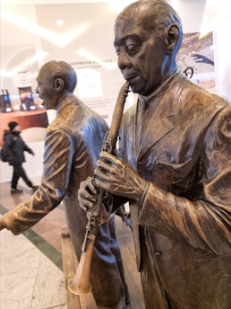 Bronze sculptures of two men in suits are depicted, with one playing a clarinet. The sculptures are highly detailed, showing textures and expressions, set in what seems to be an indoor shopping area with promotional posters in the background.