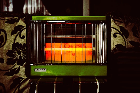 An electric heater with vertical metal bars and a glowing orange heating element is surrounded by patterned fabric upholstery. The heater has a label that reads 'LEOPET DR-622S' on a green panel.