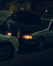 A person leans into the open hood of a car while another vehicle is nearby. It appears to be nighttime, as the scene is dimly lit with streetlights and building lights in the background.