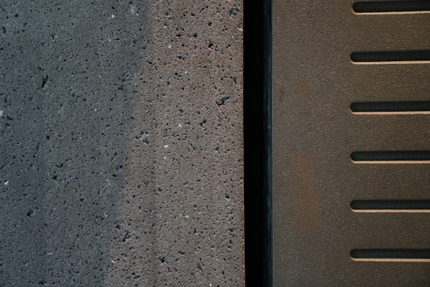 A close-up view of two adjacent surfaces, one concrete with small holes and a textured pattern, and the other metal with parallel slotted lines.