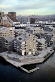 A modern urban area with distinctive, geometric residential buildings made predominantly of concrete and glass. The structures feature multiple balconies and large windows, showcasing a design that combines functionality with contemporary flair. A waterfront area and dock can be seen in the foreground, hinting at the neighborhood's integration with the water. There are a few people scattered around the area, contributing to a sense of quiet everyday activity. In the background, city skyscrapers and cranes suggest ongoing urban development.