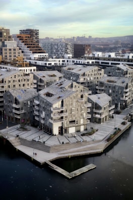 A modern urban area with distinctive, geometric residential buildings made predominantly of concrete and glass. The structures feature multiple balconies and large windows, showcasing a design that combines functionality with contemporary flair. A waterfront area and dock can be seen in the foreground, hinting at the neighborhood's integration with the water. There are a few people scattered around the area, contributing to a sense of quiet everyday activity. In the background, city skyscrapers and cranes suggest ongoing urban development.