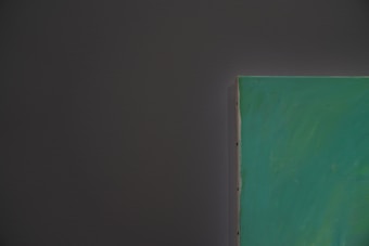 A close-up of the edge of a painting with a green canvas against a dark gray wall. The painting is partially visible, and the texture of the wall and canvas can be noted.