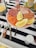 A plate of assorted deli meats and lemon wedges is placed on a striped black and white tablecloth. The plate contains sliced salami, prosciutto, ham, smoked salmon, and several small sausages. Beside the plate, there is a fork with a wooden handle labeled 'Goat' and another ornate fork. A bowl with shredded cheese and a mustard jar are also visible.