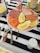 A plate of assorted deli meats and lemon wedges is placed on a striped black and white tablecloth. The plate contains sliced salami, prosciutto, ham, smoked salmon, and several small sausages. Beside the plate, there is a fork with a wooden handle labeled 'Goat' and another ornate fork. A bowl with shredded cheese and a mustard jar are also visible.