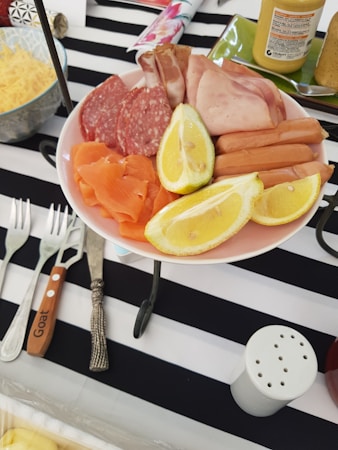 A plate of assorted deli meats and lemon wedges is placed on a striped black and white tablecloth. The plate contains sliced salami, prosciutto, ham, smoked salmon, and several small sausages. Beside the plate, there is a fork with a wooden handle labeled 'Goat' and another ornate fork. A bowl with shredded cheese and a mustard jar are also visible.